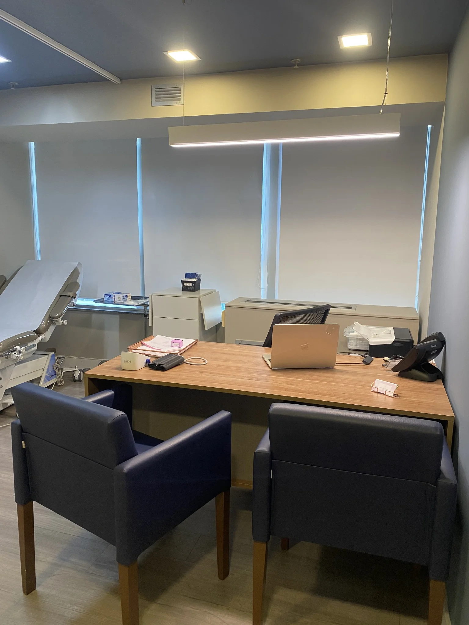 Mesa de atendimento do consultório com duas poltronas azuis, notebook e maca ginecológica ao fundo