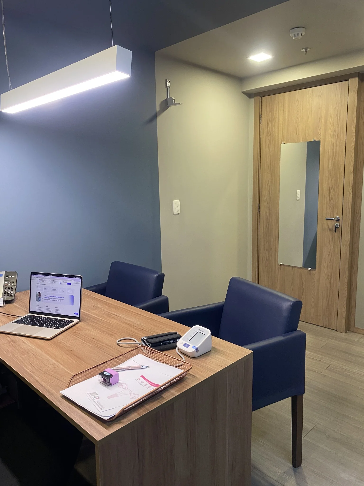 Mesa de atendimento com notebook, poltronas azuis e porta de madeira com espelho
